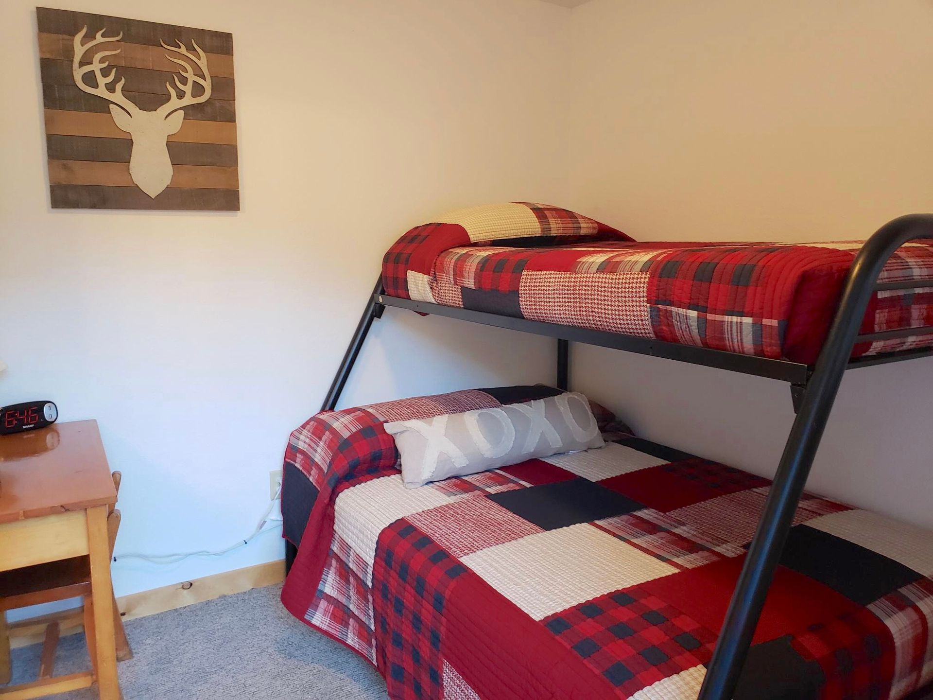 Bunk beds in a room, red plaid bedding, a wooden deer head wall decor, and a small desk.