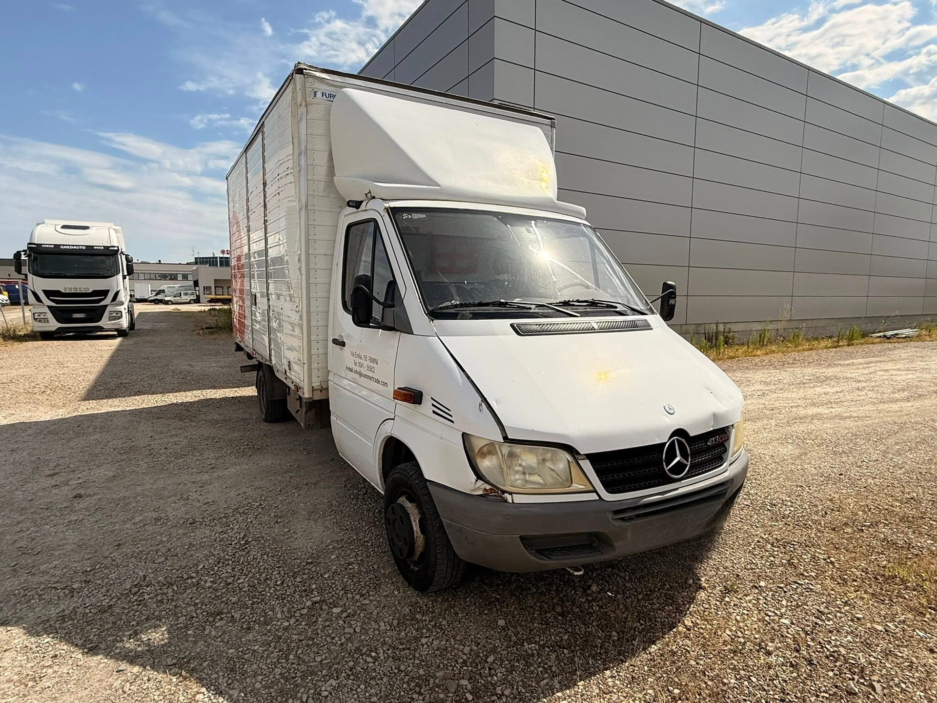 Un furgone bianco è parcheggiato in uno spiazzo ghiaioso di fronte a un edificio.