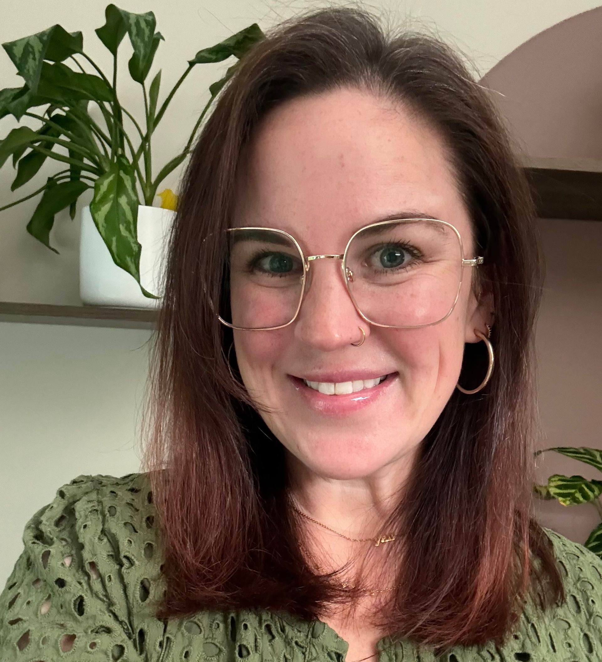 Smiling woman with glasses and hoop earrings in a green top, standing indoors near plants.