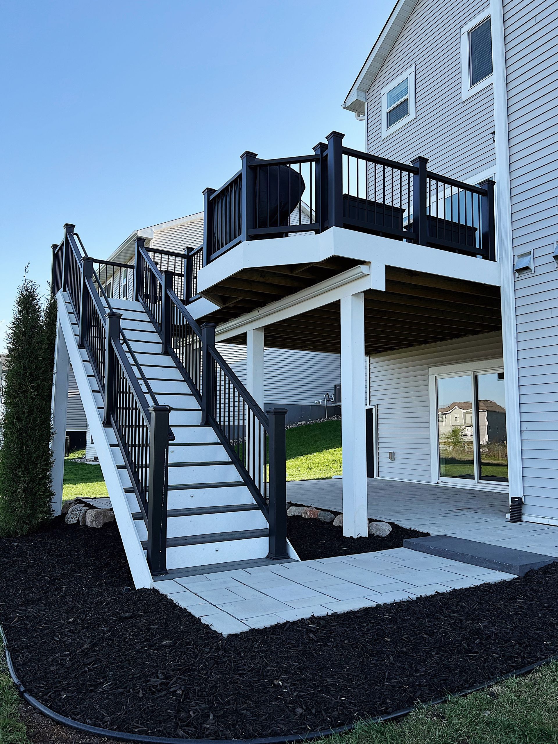 A two-story deck with black railings and stairs. The deck is attached to a white house with a paved patio below.