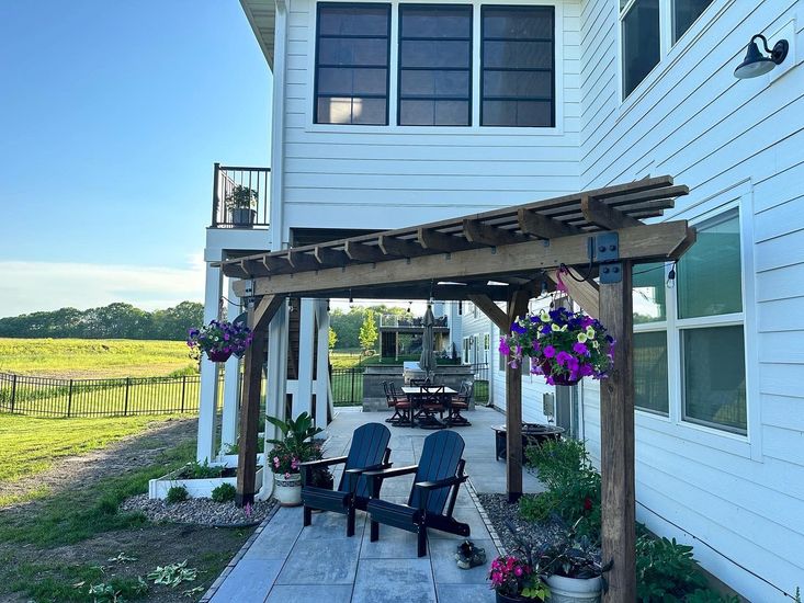 Outdoor patio with pergola, Adirondack chairs, and hanging flowers, next to a white house.