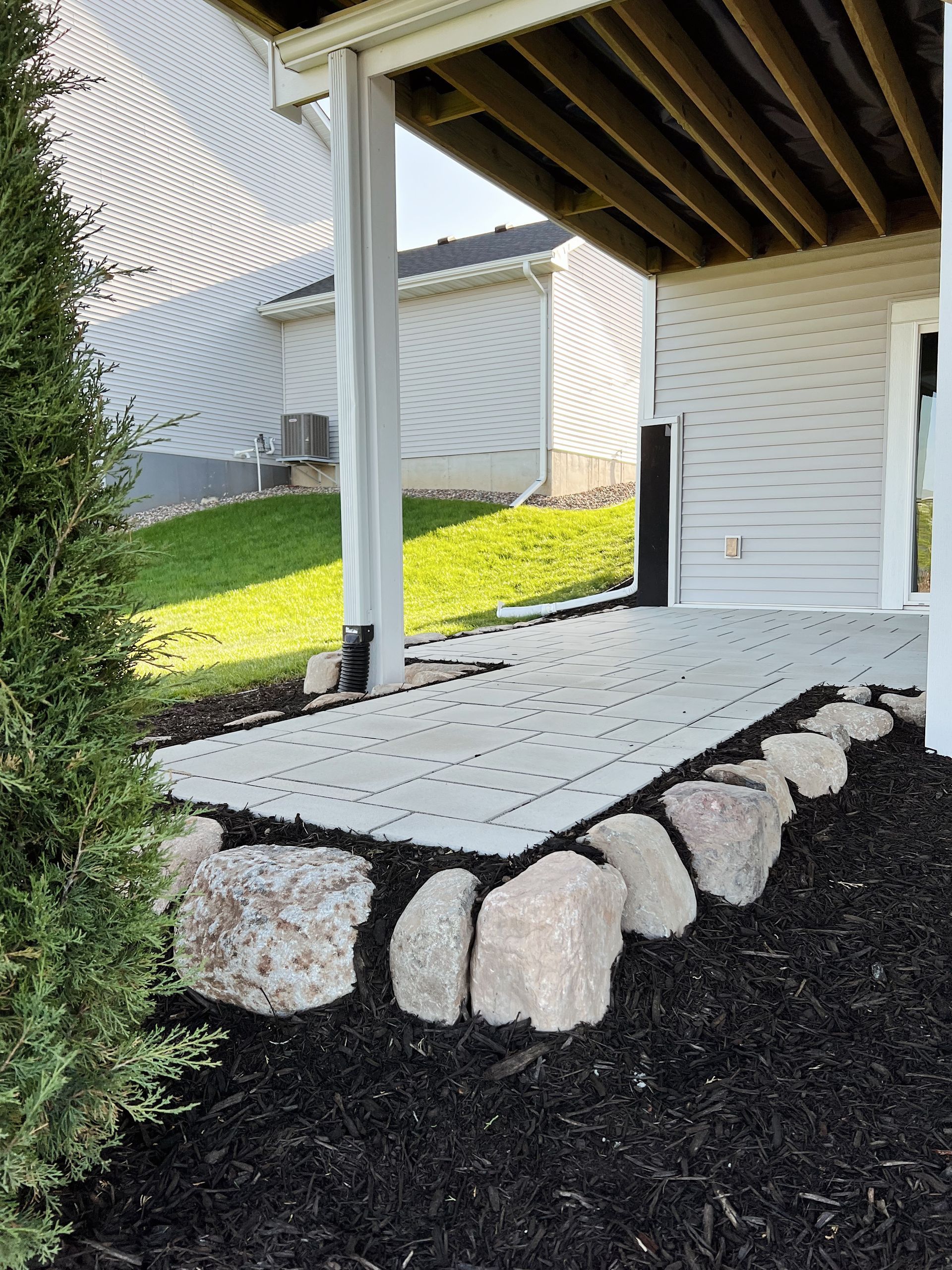 Stone patio with large rocks, black mulch, and a green lawn under a deck.