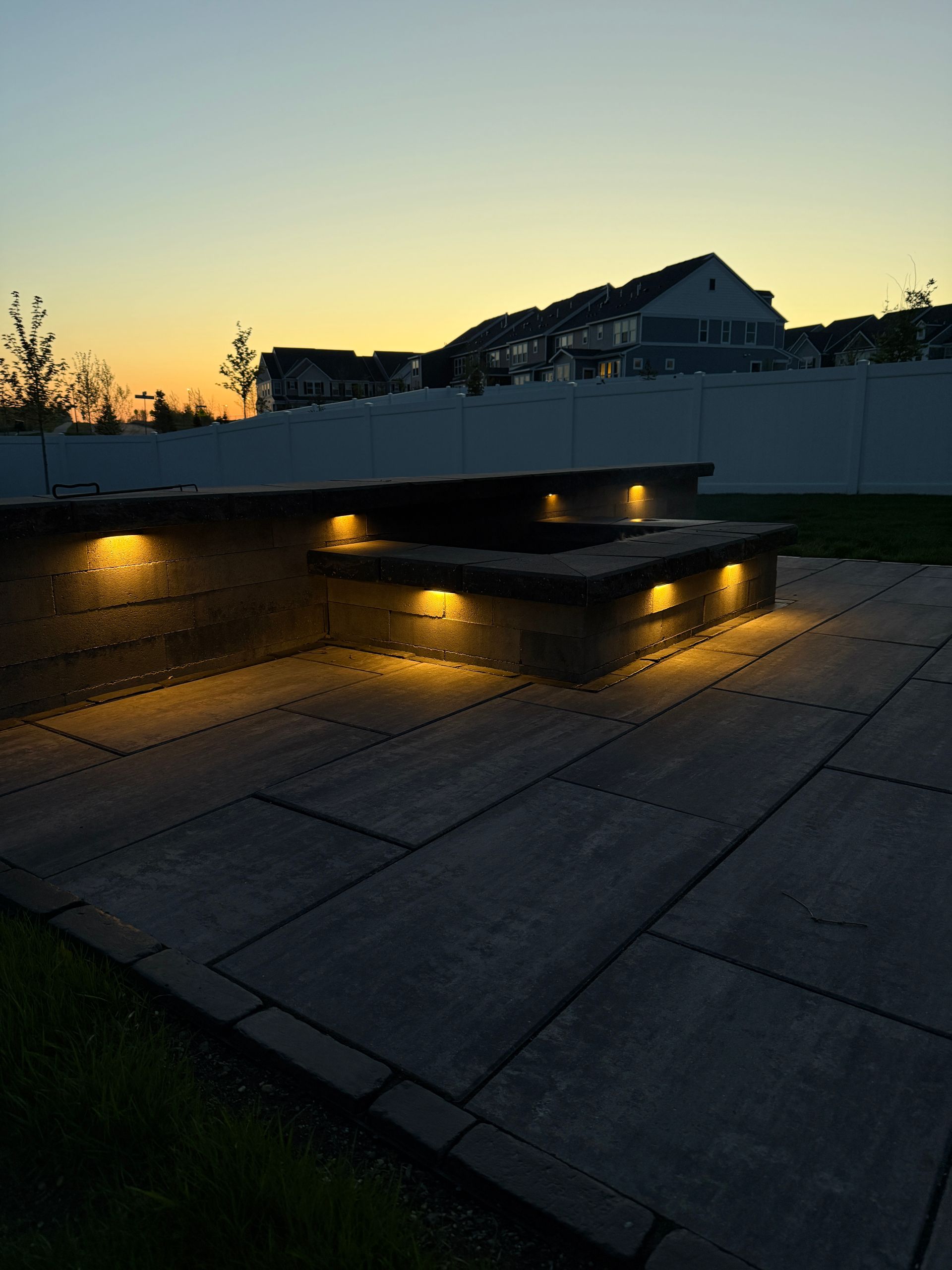 A tiered, illuminated patio seating area at dusk; residential houses in the background.