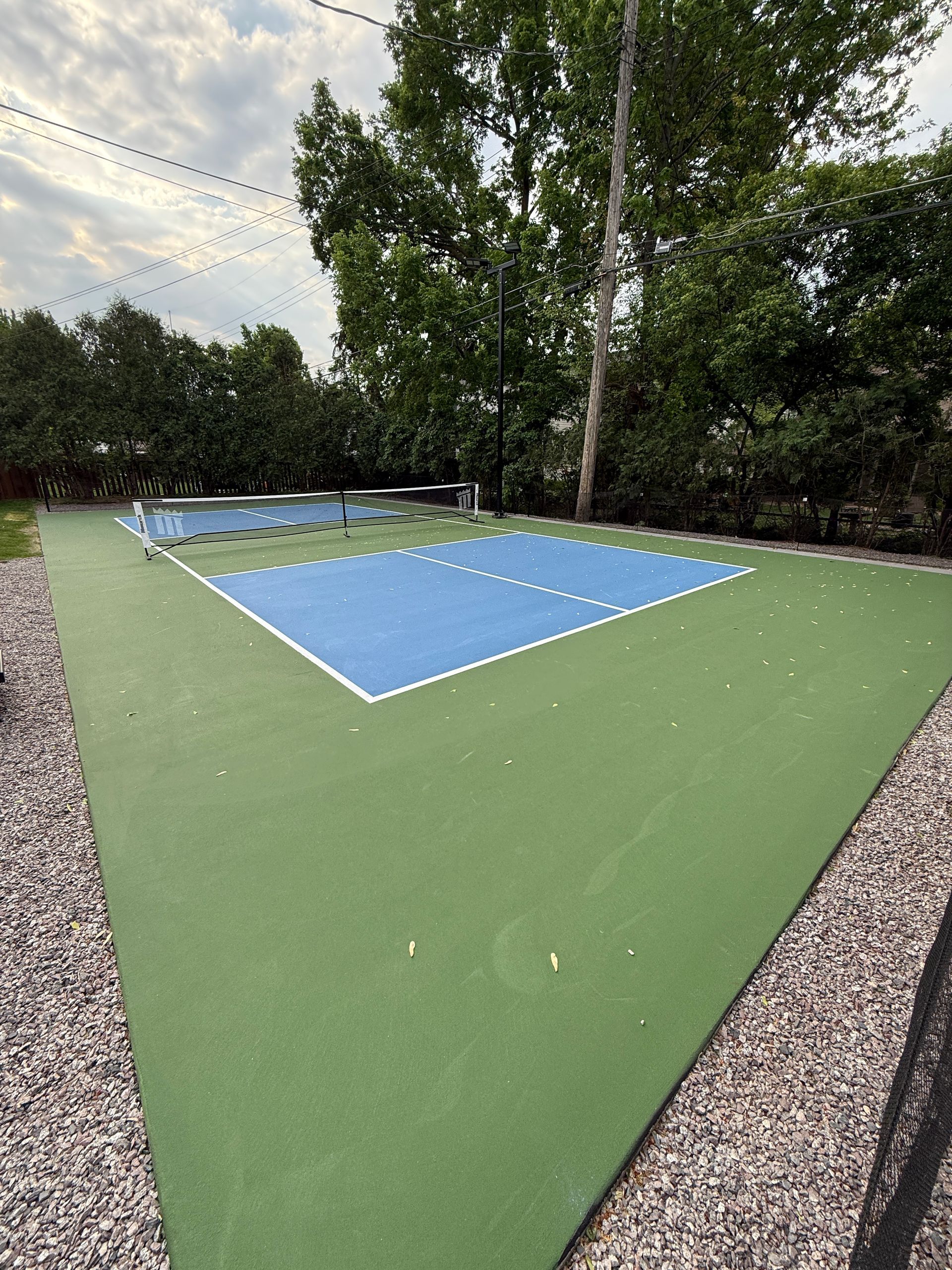 Outdoor pickleball court with blue playing area, green surrounding surface, and gravel border.