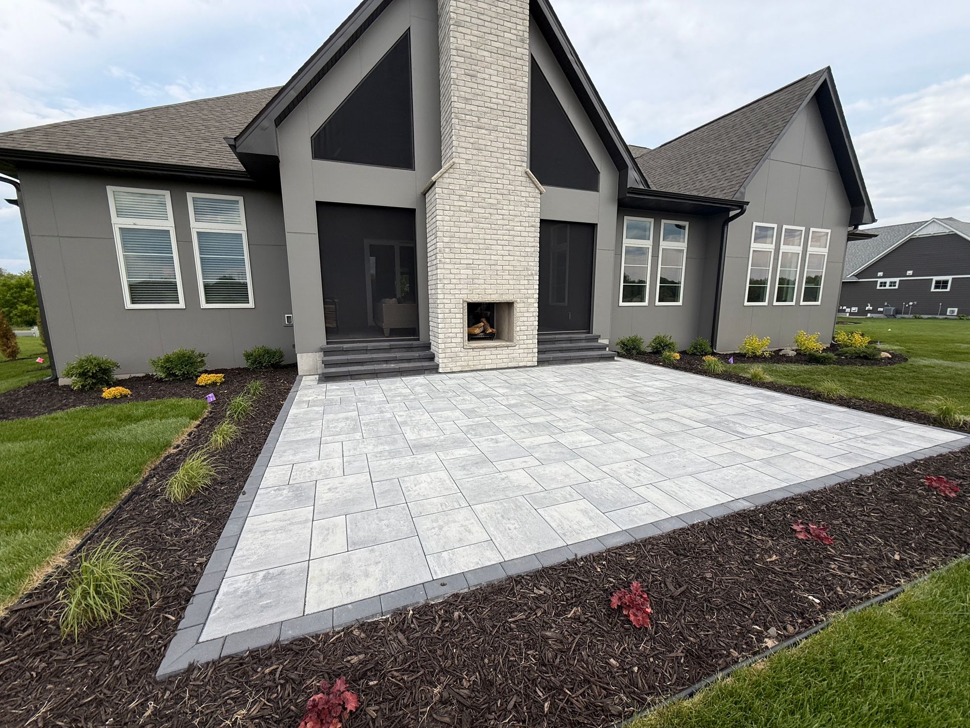 Backyard patio with gray pavers, built-in fireplace, and landscaping in front of a house.