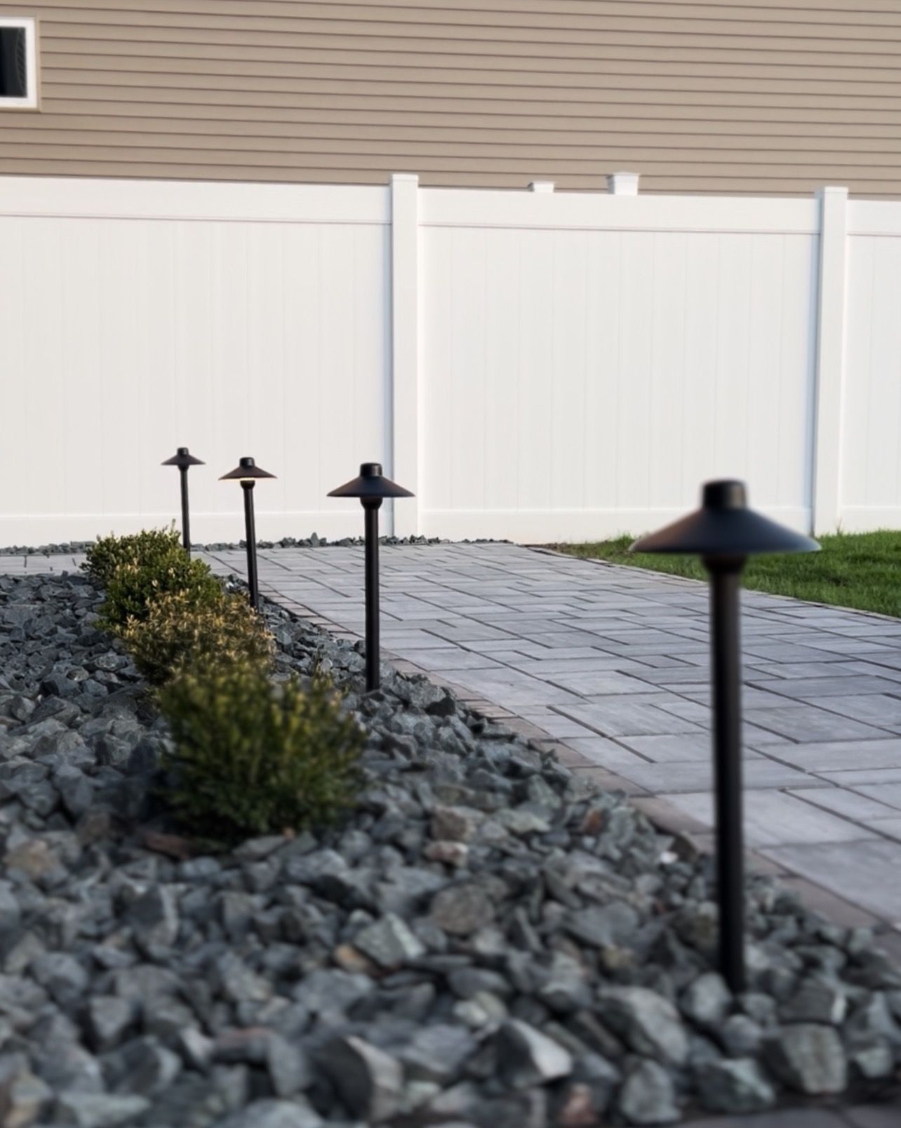 Path with landscape lights and shrubs next to a white fence.