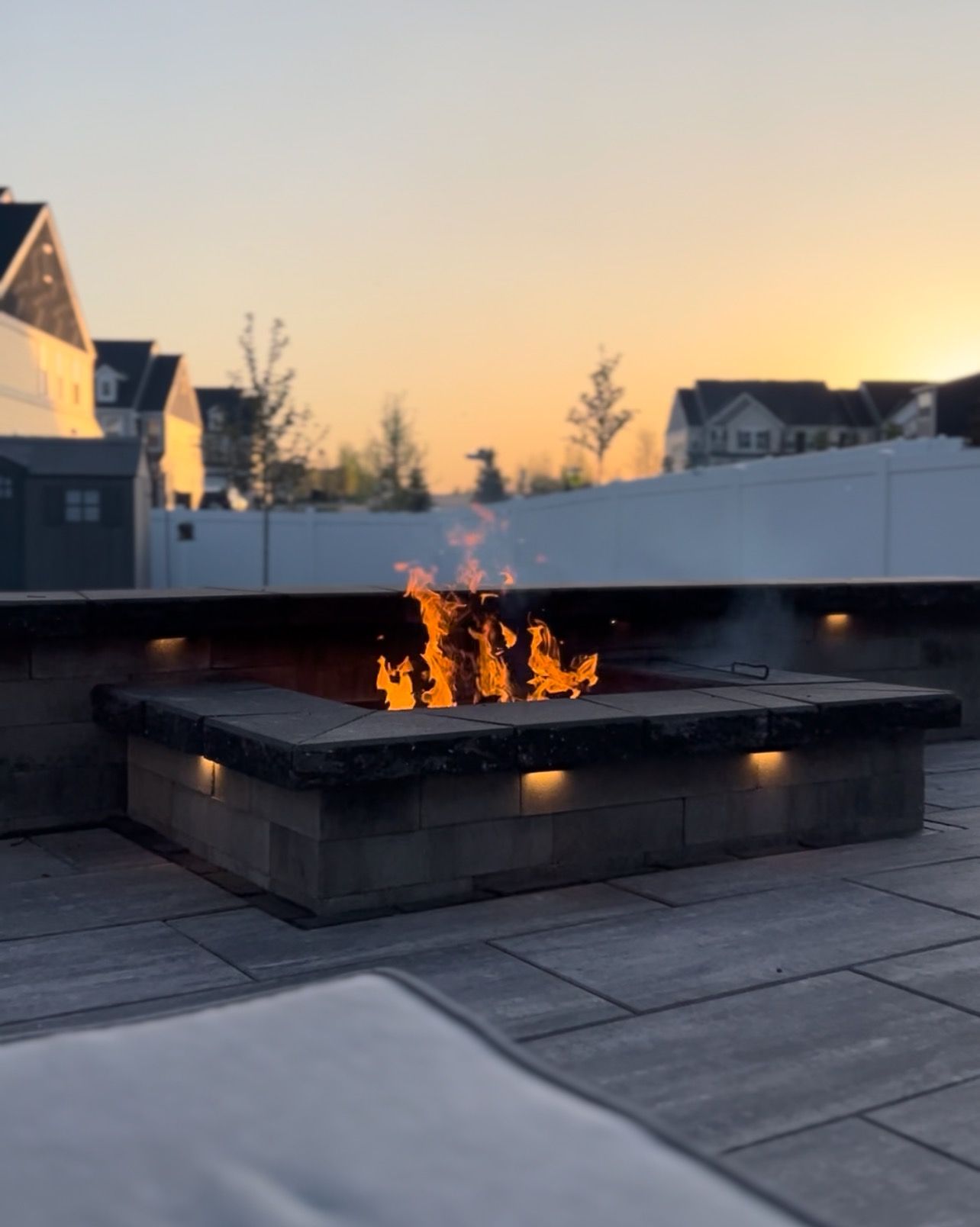 Fire pit with flames, lit at dusk. Stone pavers, ambient lighting, and suburban backdrop.