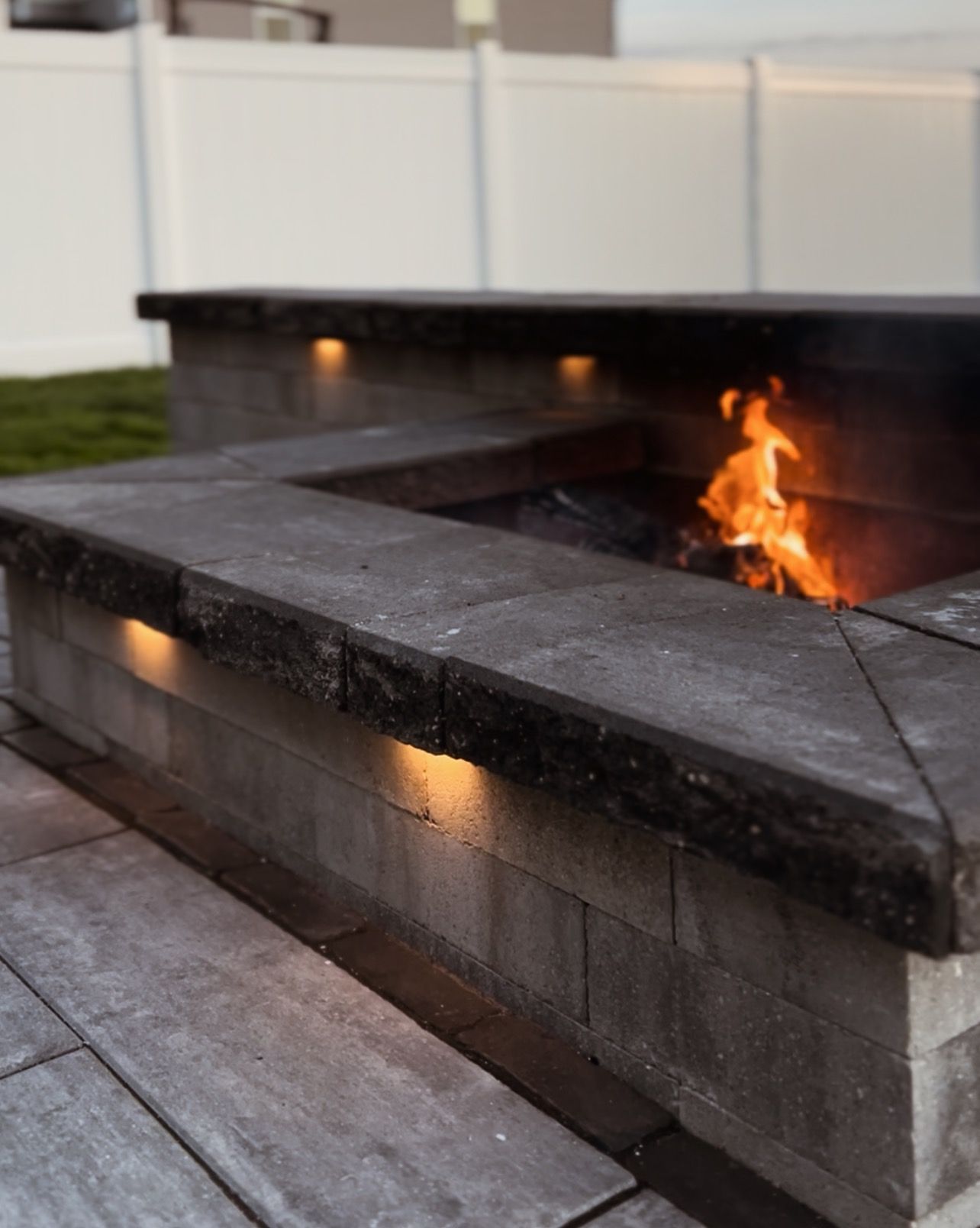 Stone fire pit with built-in lights, burning fire, set on a stone patio with white fence in the background.