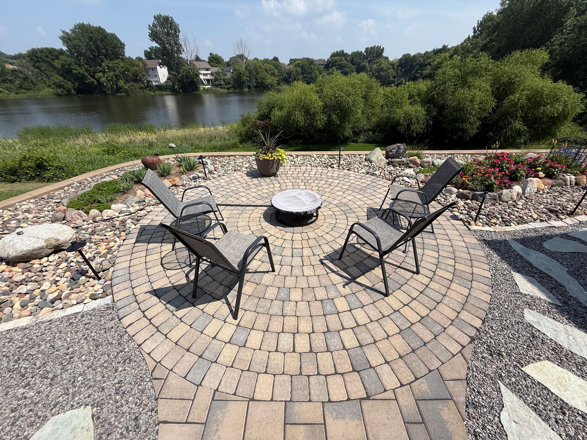 Circular brick patio with four chairs around a fire pit, overlooking a river with green trees.