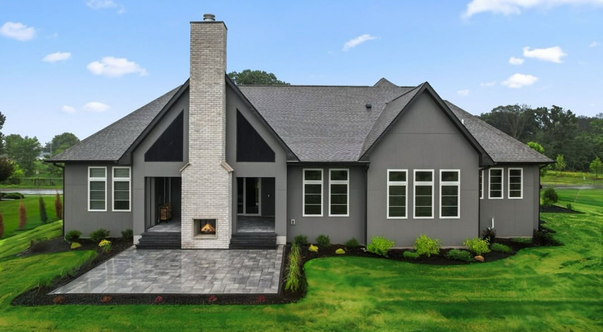 Grey house with stone chimney, patio, and green lawn under a blue sky.
