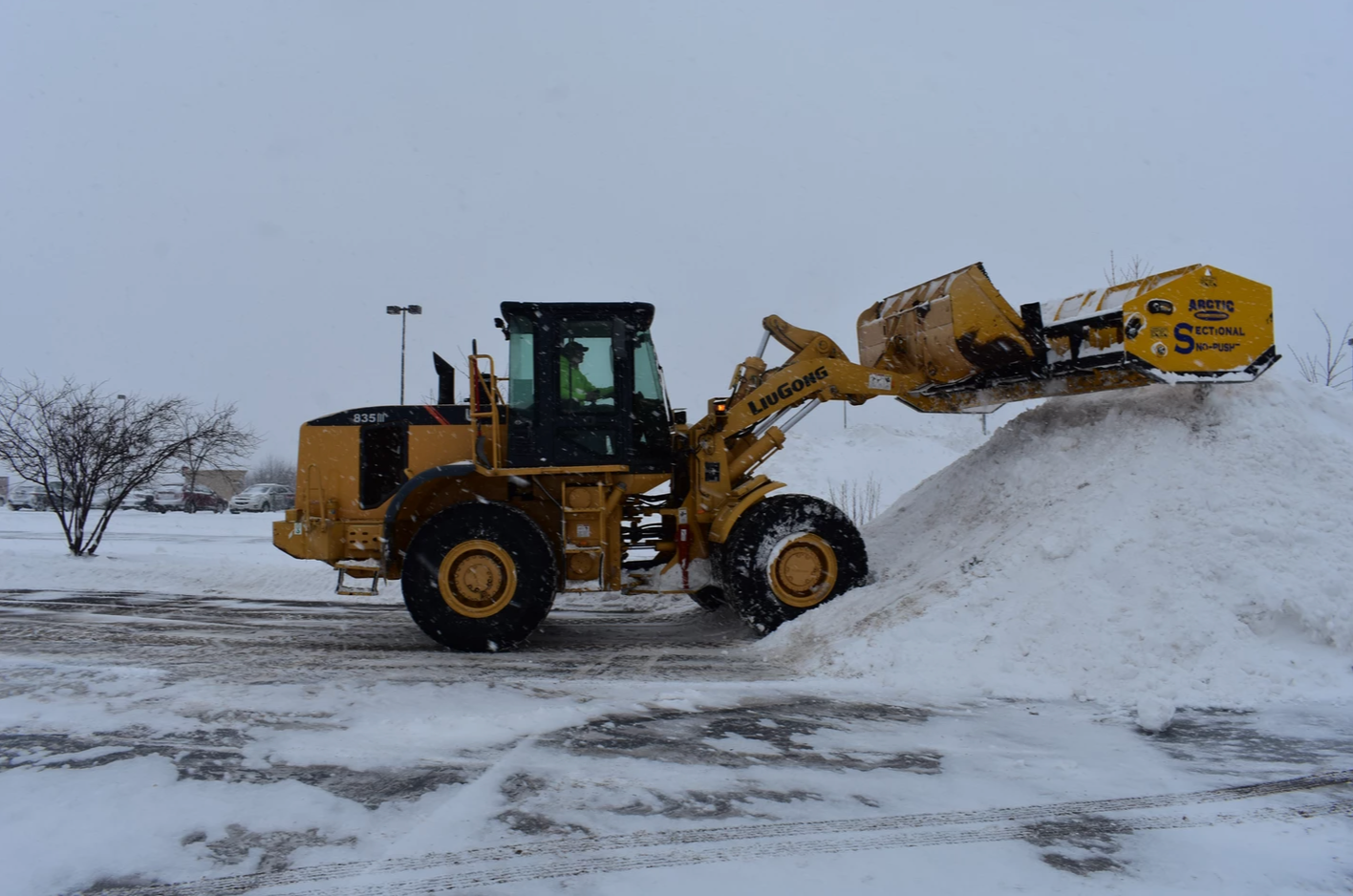 Yellow loader plowing snow in a parking lot on an overcast day.
