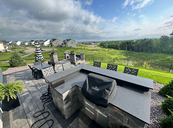 Outdoor kitchen with a grill, overlooking a green field and a residential neighborhood under a cloudy sky.