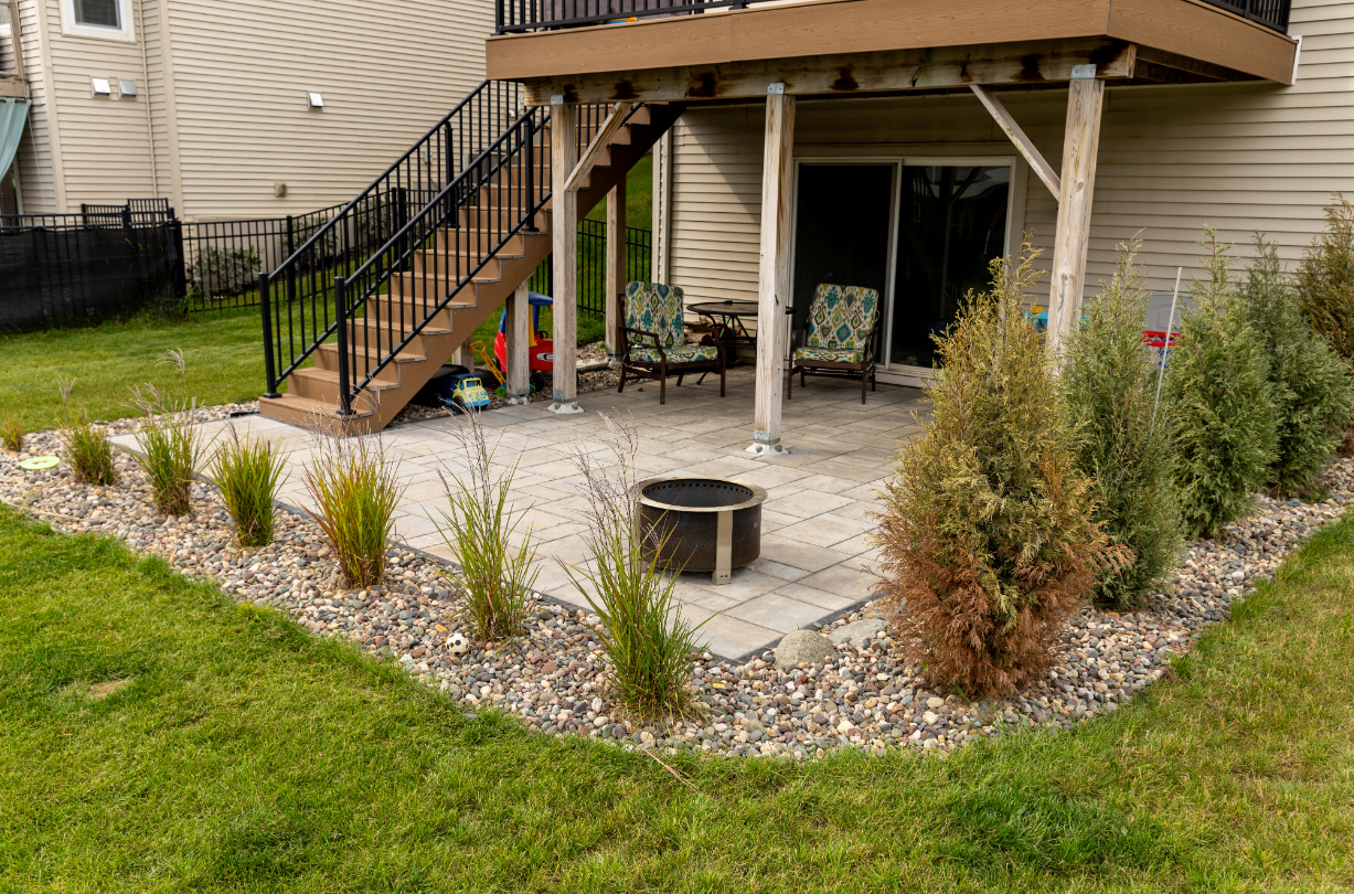 Backyard patio with fire pit, under a deck with stairs, surrounded by landscaping and lawn.