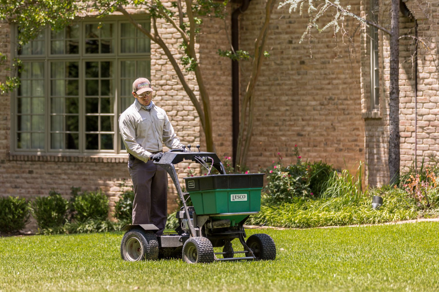 Man using a green fertilizer spreader on a lawn in front of a brick building.