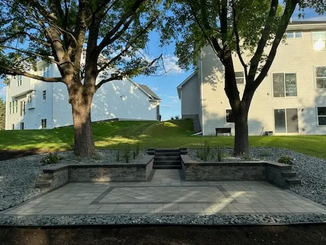 Stone patio with trees, retaining walls, steps, and houses in the background on a sunny day.
