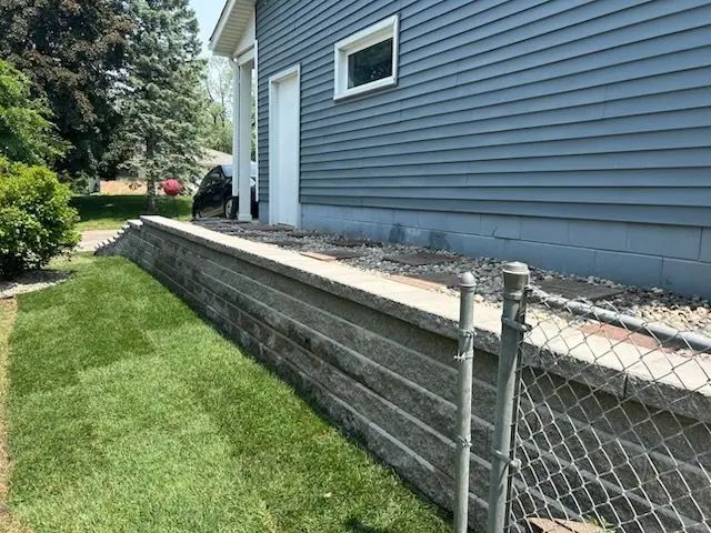 Gray retaining wall beside a house with blue siding and a chain-link fence on a grassy lawn.