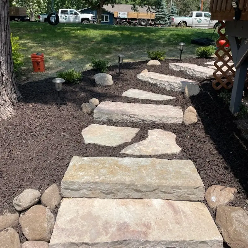 Stone pathway through dark mulch, flanked by rocks and small lights in a landscaped yard.