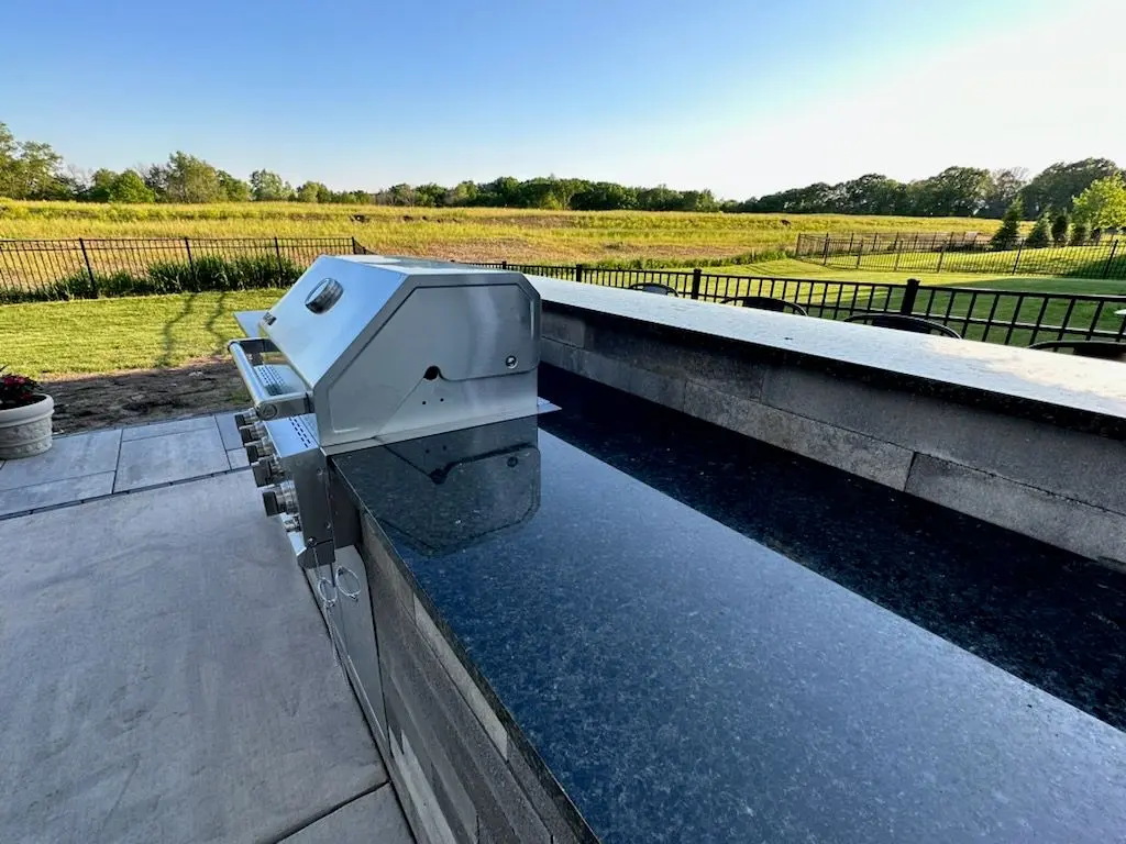 Outdoor kitchen with stainless steel grill, black granite countertop, and grassy field background.