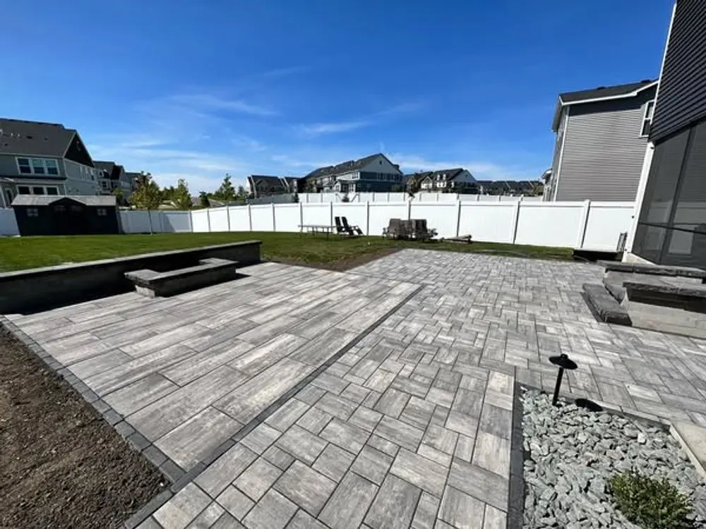 Backyard patio with gray pavers, grass lawn, white fence, and blue sky.