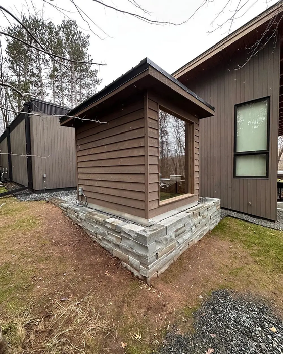 Brown shed-like structure with a large window, on a stone foundation, next to a modern building.