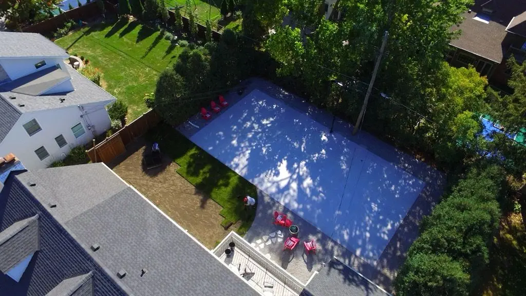 Aerial view of a rectangular pool surrounded by a patio and lawn, with trees and houses visible.