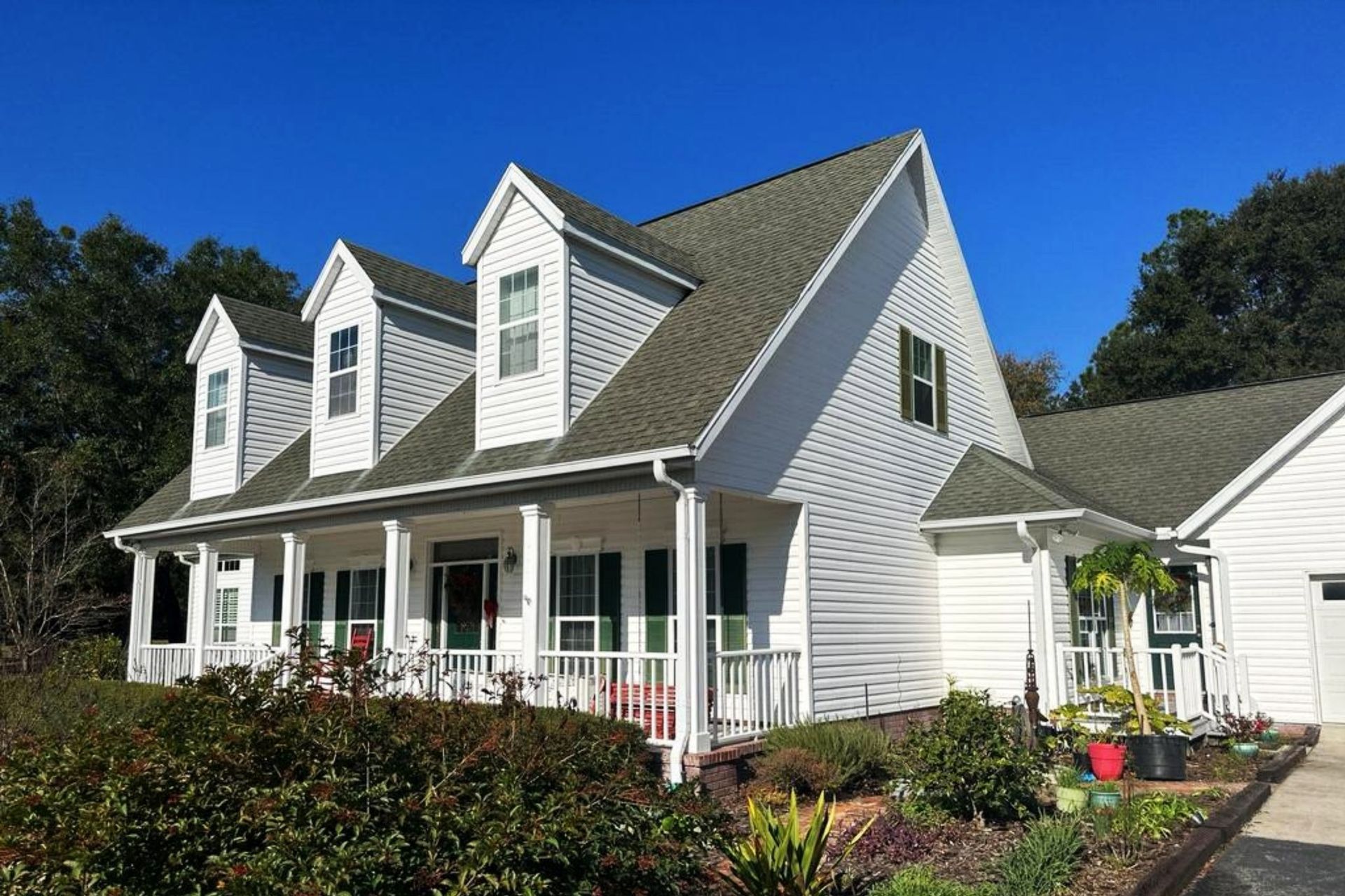 A white house with an open porch, and a clean, new roof. It is made of gray shingles. There is a green garden surrounding the house.