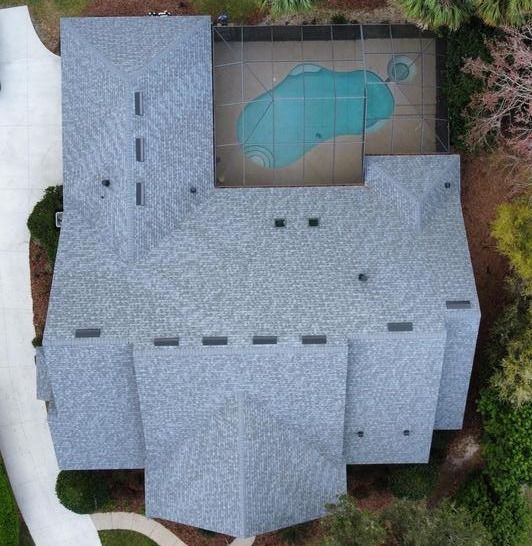 An aerial view of the top of a residential house. The main view is the clean, new roof of the house, made of gray shingles. There is a backyard at the end corner of the house with a big abstract-shape pool. Surrounding the house there is a long parkway at the left, and a forest at the right.