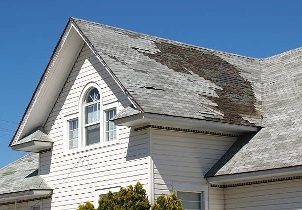 A white residential property with a damaged gray shingles' roof with fallen shingles on the middle of the roof.