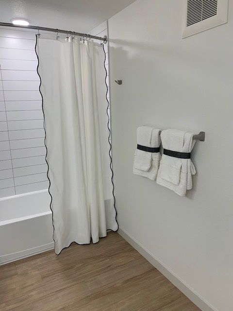 A modern bathroom with a white shower curtain, light wood flooring, and two white towels hanging on a wall rack.