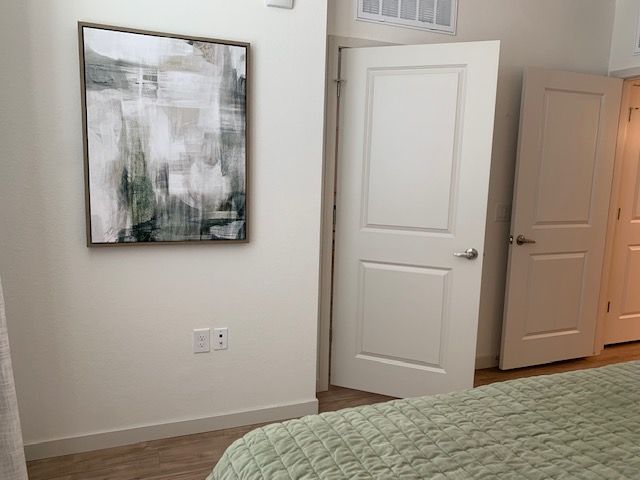 An abstract painting with green and white tones hangs on a white wall above a bedroom corner with two open doors.