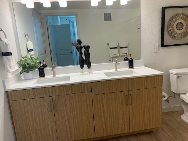 A modern double-sink bathroom vanity with light wood cabinets, white countertops, a large mirror, and hanging wall art.