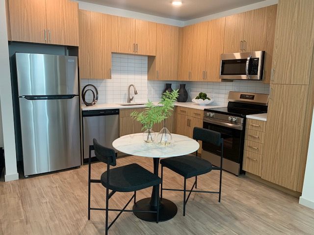 A modern kitchen with light wood cabinets, stainless steel appliances, and a small round dining table with two chairs.