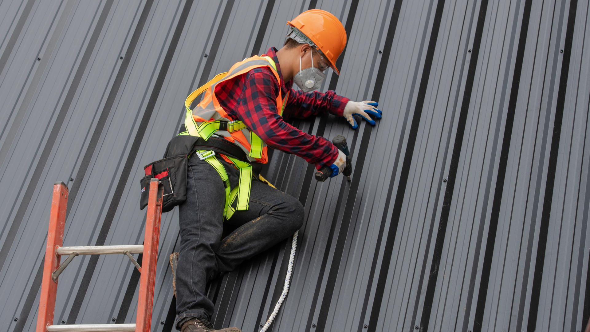 Worker on ladder, installing metal roof; wearing helmet, harness, safety vest, and respirator.