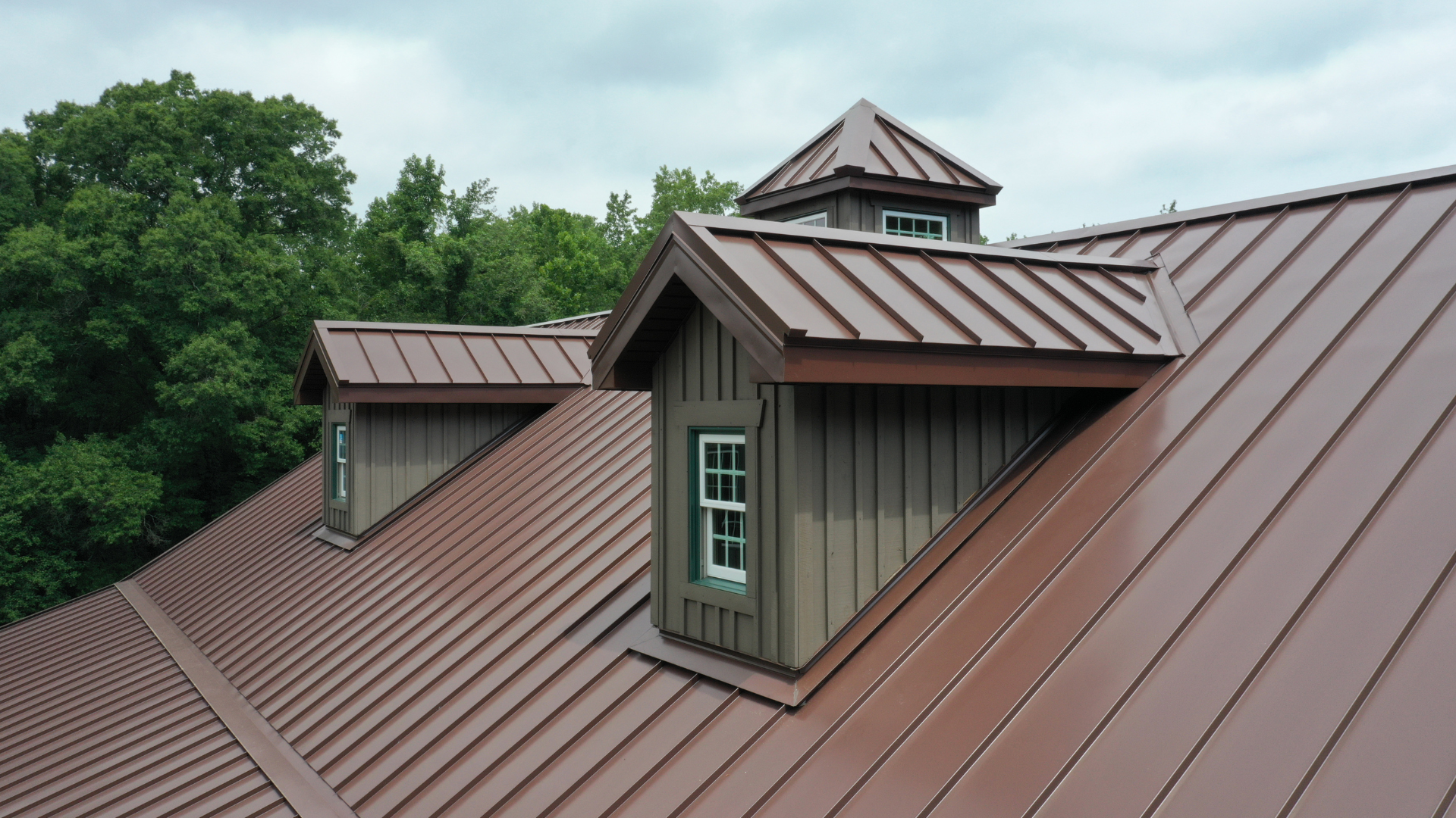 Brown metal roof with dormers, set against a green tree backdrop and cloudy sky.