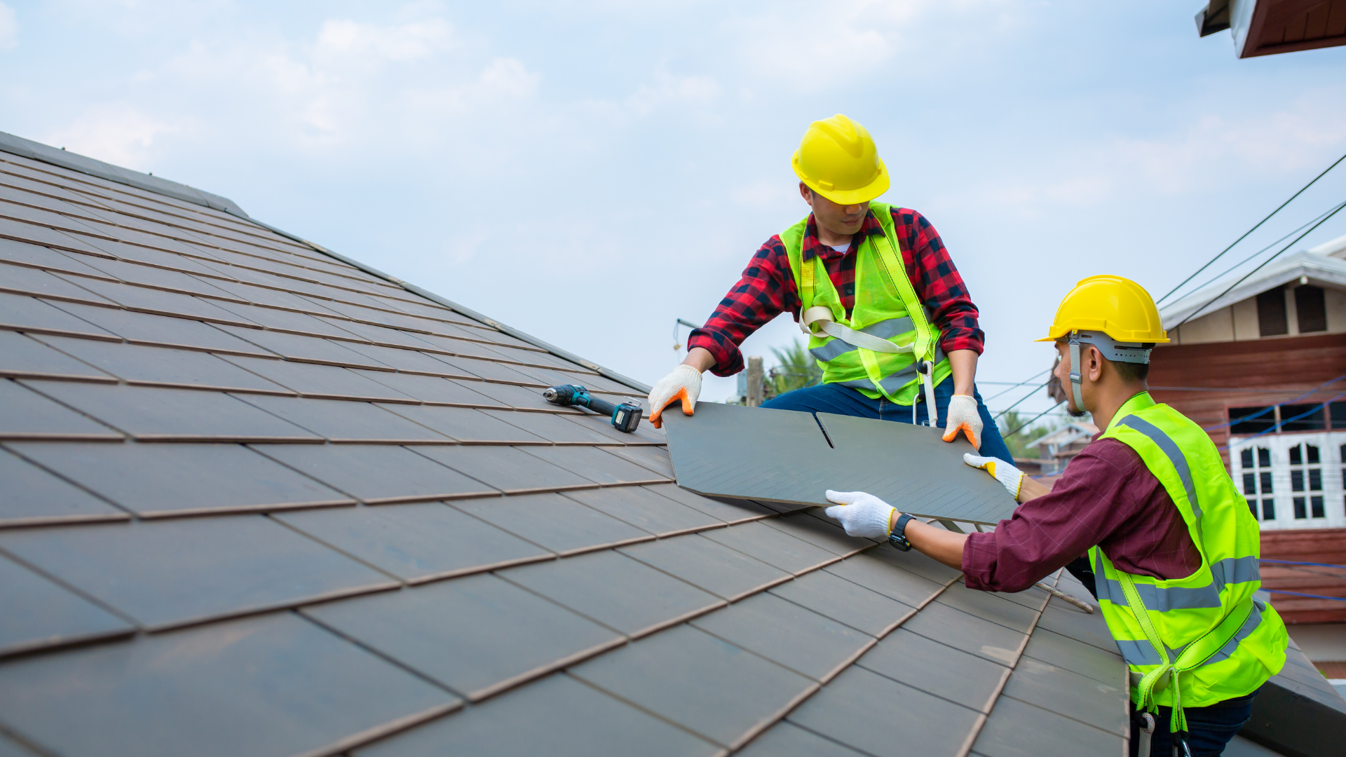 Two roofers installing dark gray shingles on a house roof, wearing safety vests and hard hats.