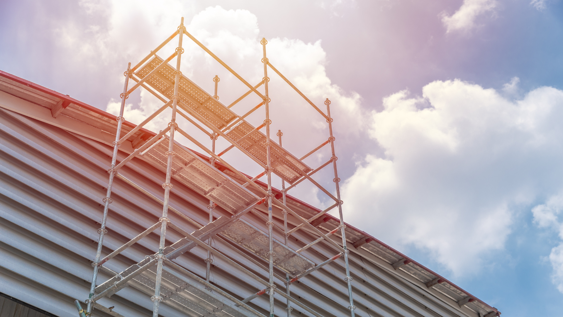 Scaffolding on a corrugated metal roof against a bright blue sky with puffy white clouds.