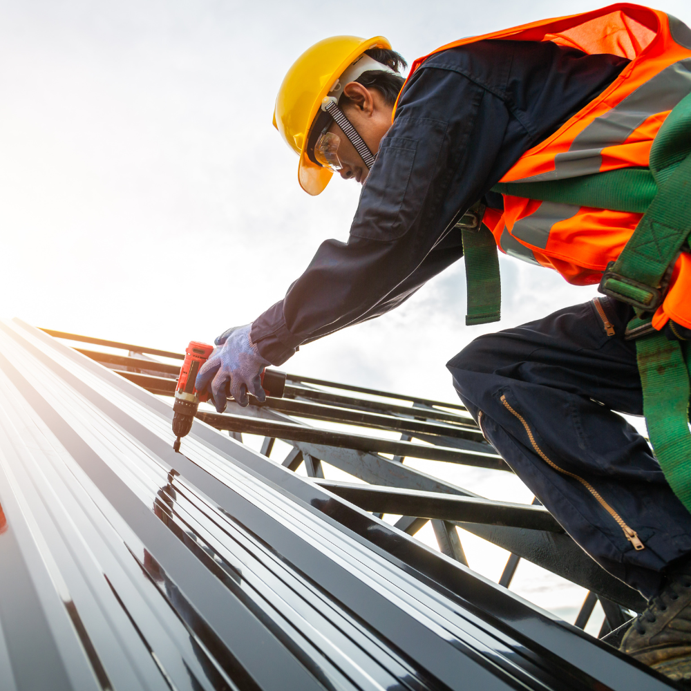 Roofer in safety gear installing metal roofing with a power drill on a sunny day.