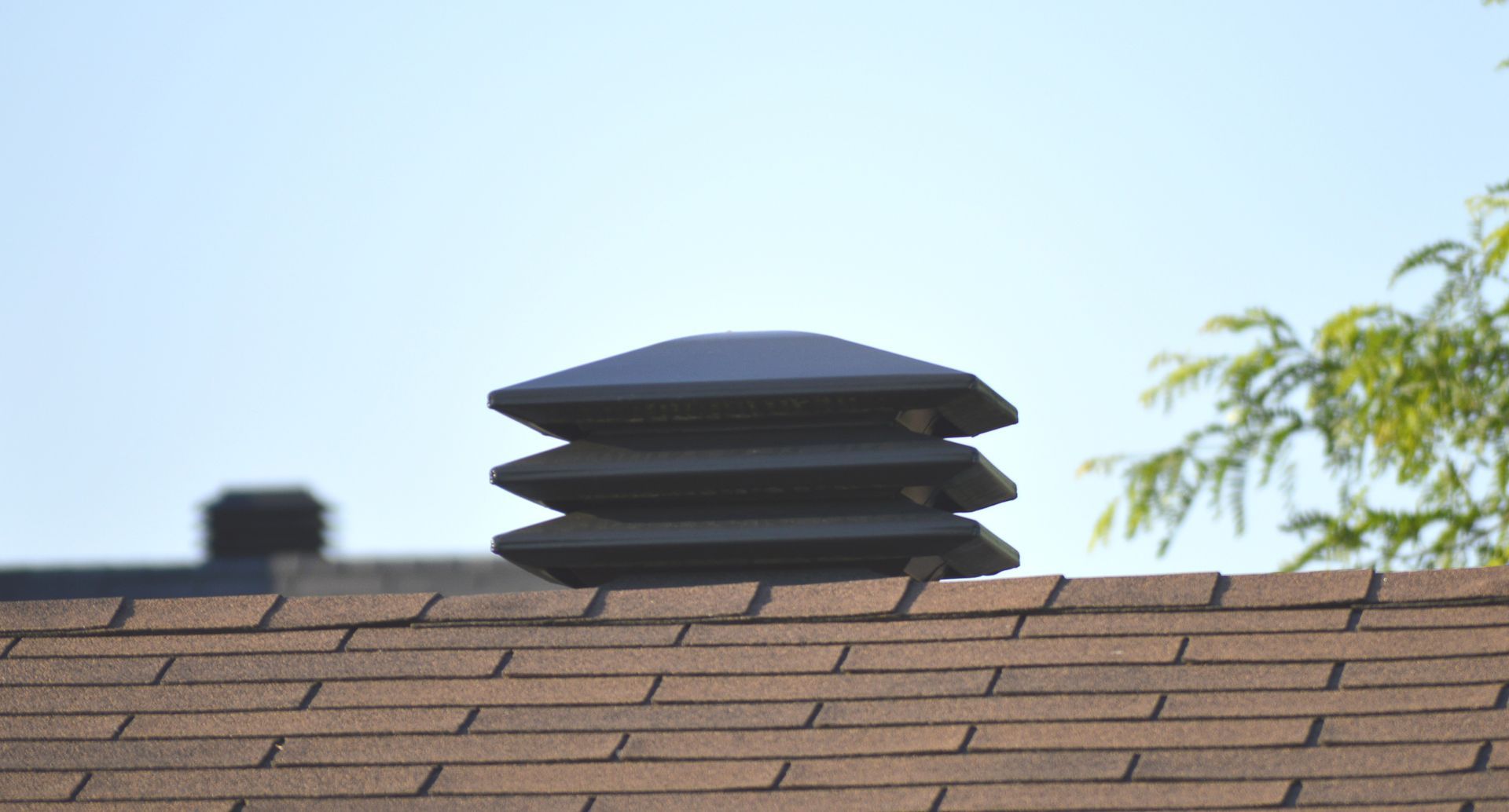 Dark, tiered roof vent on a brown shingled roof against a blue sky.