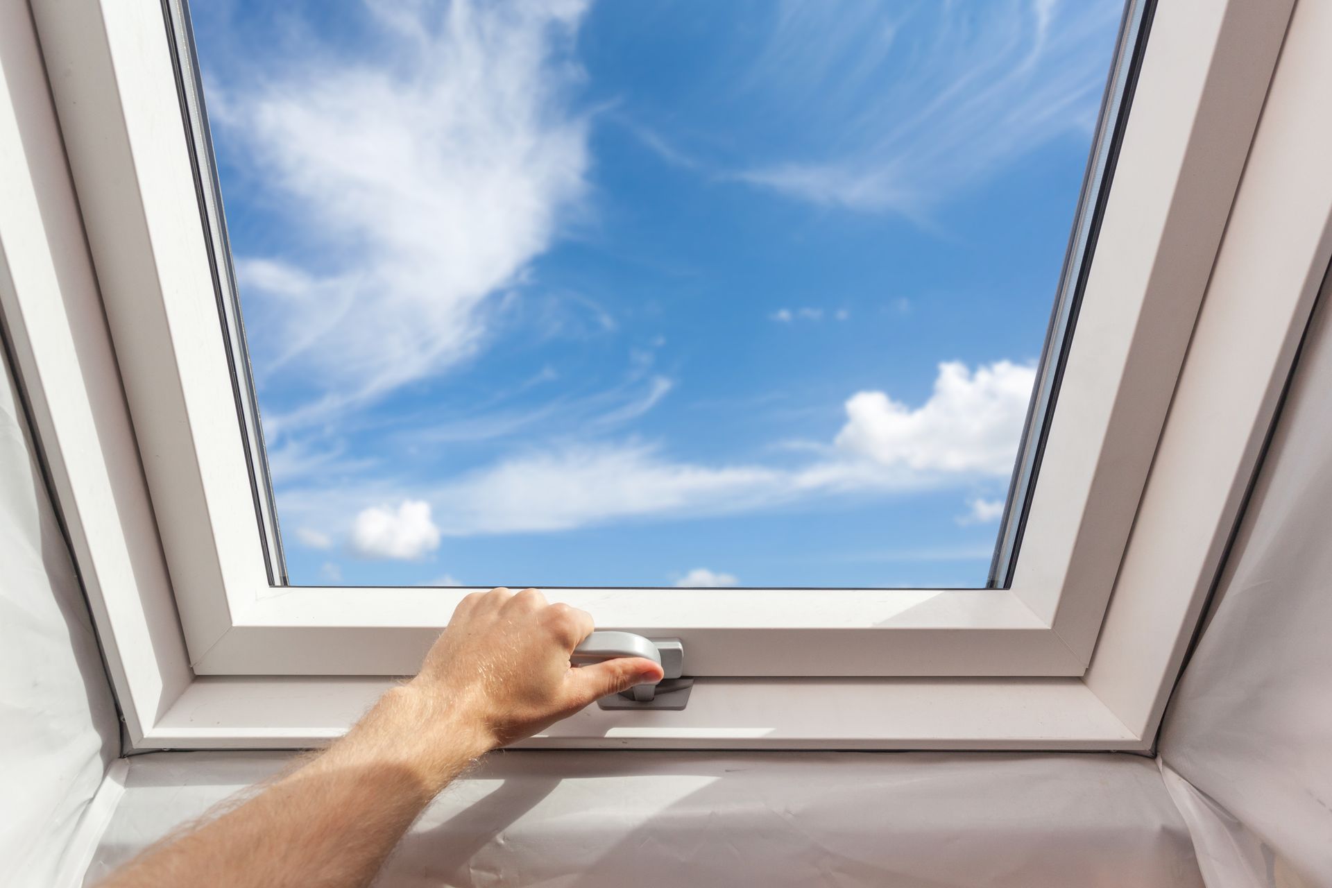 Hand opening a white-framed skylight, revealing a bright blue sky with wispy clouds.
