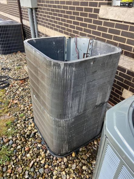 Damaged air conditioner coil unit with exposed fins, against a brick wall, next to another unit and gravel.