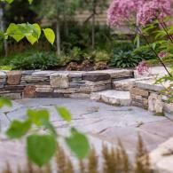 Stone garden patio with curved bench, greenery, and pink flowers in the background