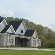 Modern gray house with white trim and peaked roofs beside a grassy yard and wooded hillside