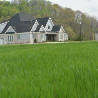 Large white house with dark roof behind a green grassy field and trees