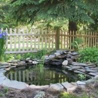 Stone-lined garden pond beneath a wooden fence and leafy trees