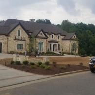 Large stone house with a dark roof and landscaped front yard along a quiet street