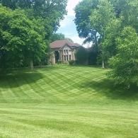 Large house atop a manicured green hill, framed by dense trees and a bright blue sky