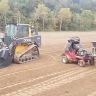 Blue bulldozer and red lawn tractor on a dirt field with trees in the background