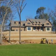 Large beige house on a hillside with a green lawn, bare trees, and construction equipment in front