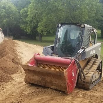 Red skid steer loader pushing sand along a dirt path beside trees