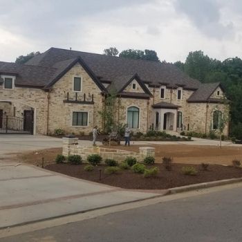 Stone house with dark roof and arched entry, front landscaping, and a person walking in the driveway